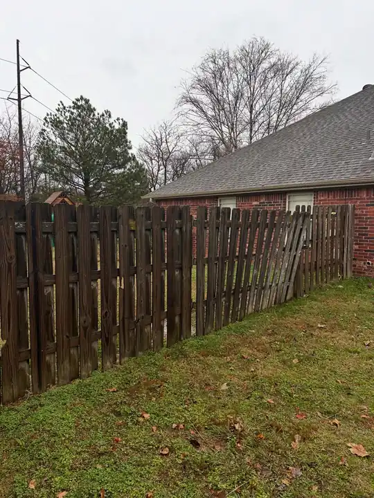 An older weathered spaced-picket fence behind a brick house, the kind of run that comes in for repair