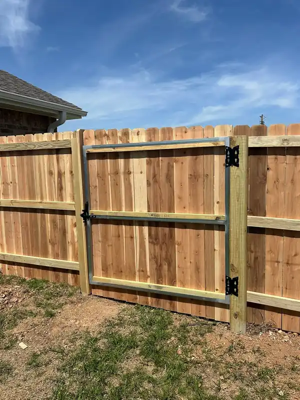 Dog-ear cedar fence with a galvanized steel-framed walk gate and black hinges, house eave at left