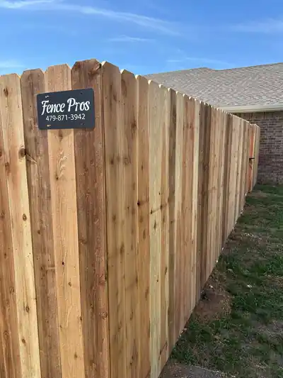 Dog-ear cedar privacy fence beside a brick house corner, with the Fence Pros company sign on the fence